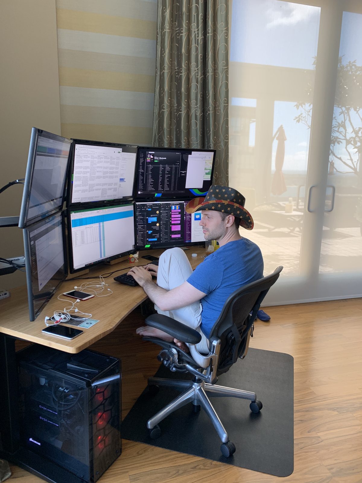 Adam Young in an American flag cowboy hat working barefoot at a six-monitor battlestation