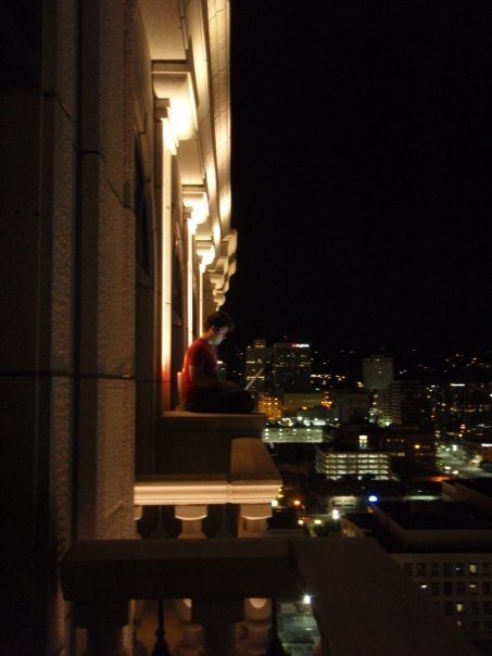 A young Adam Young sits with a laptop on a high-rise ledge at night