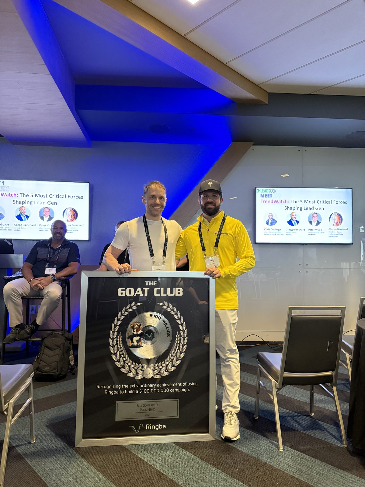 Adam Young and Bill Somerville of PolicyBind holding a giant $100 million GOAT Club plaque