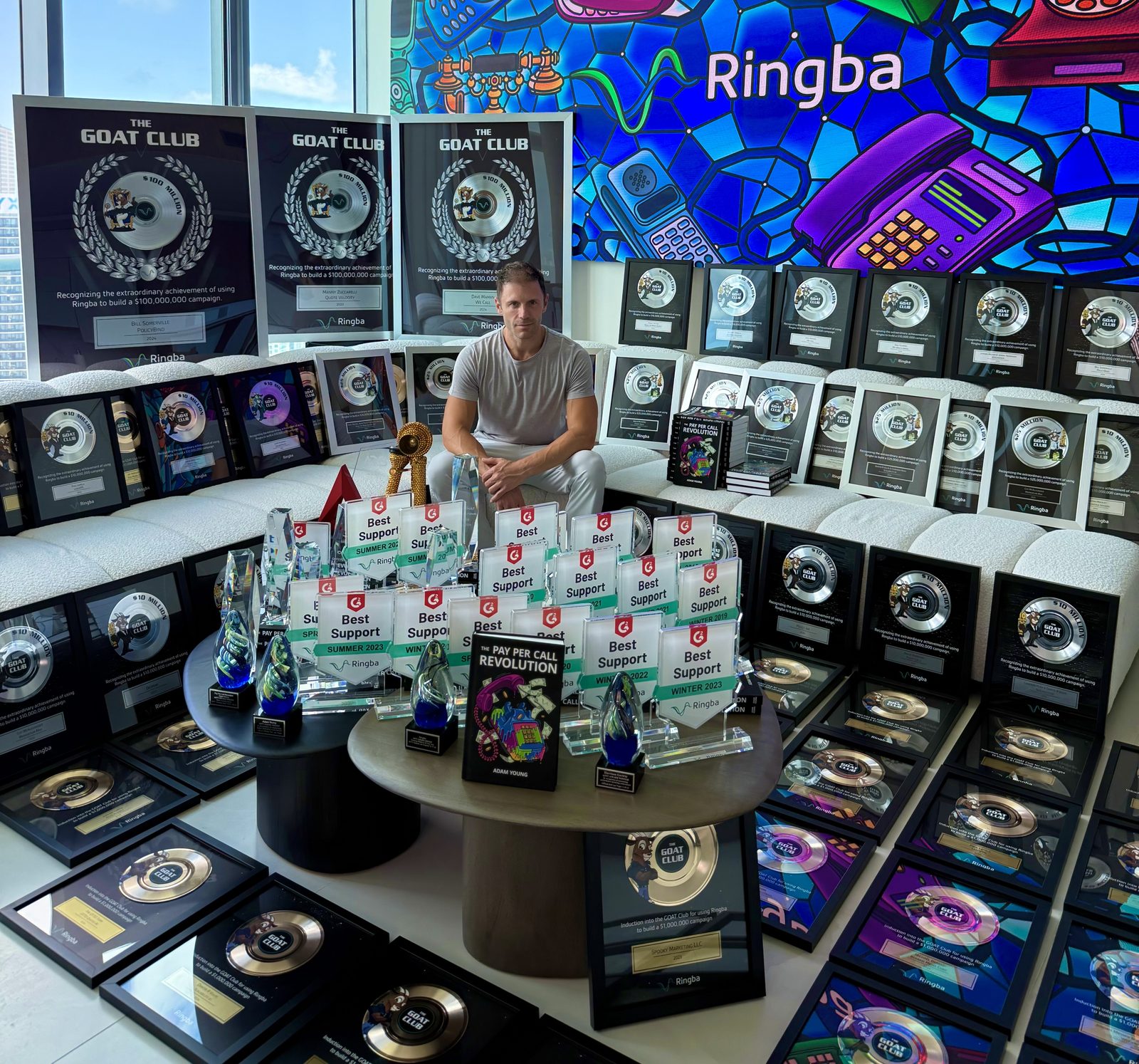 Adam Young seated among GOAT Club plaques, awards and his books beneath a stained-glass Ringba mural