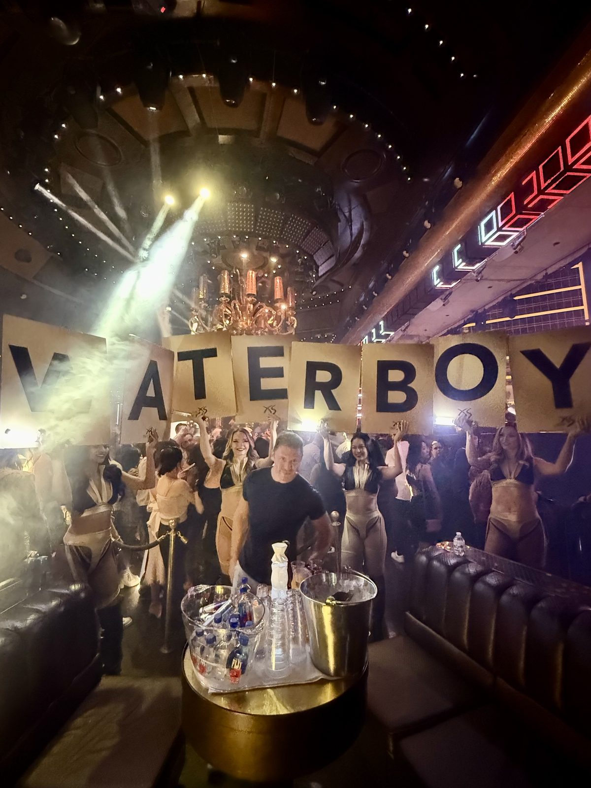 Dancers hold letter cards spelling WATERBOY over Adam Young's table stacked with water bottles at XS Las Vegas